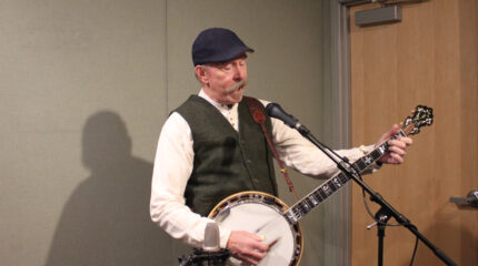 An adult playing banjo and singing into a microphone in a radio studio