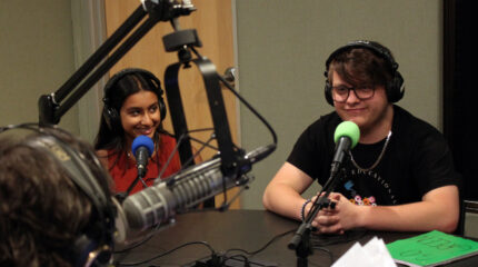 Two teens wearing headphones speaking into microphones being interviewed by an adult wearing headphones sitting behind a microphone in a radio studio