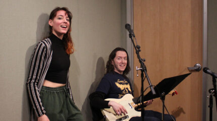 Two adults smiling at the camera, one sitting and holding an electric guitar in a radio studio