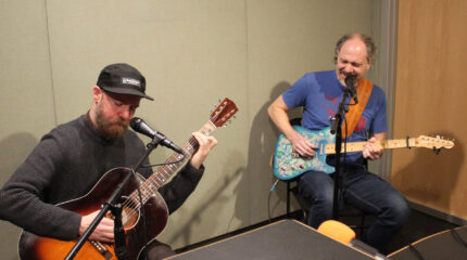 An adult playing acoustic guitar sitting behind a microphone and an adult playing electric guitar singing into a microphone in a radio studio