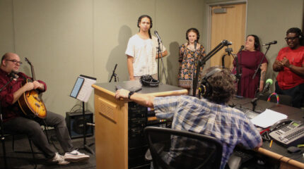 An adult sitting and holding an acoustic guitar and two teens and two adults wearing headphones and standing behind microphones being interviewed by an adult wearing headphones sitting behind a microphone and a mixing board in a radio studio