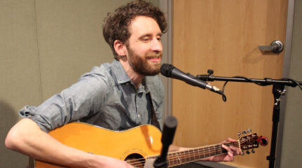 An adult playing acoustic guitar and singing into a microphone in a radio studio