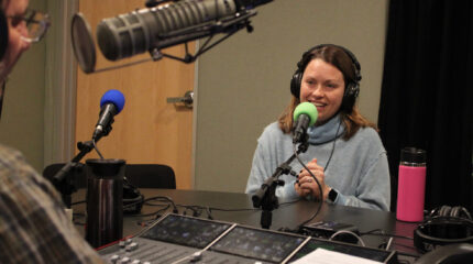 An adult wearing headphones speaking into a microphone and gesturing with their hands being interviewed by an adult wearing headphones sitting behind a microphone and a mixing board in a radio studio
