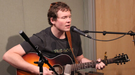 An adult playing acoustic guitar and singing into a microphone in a radio studio