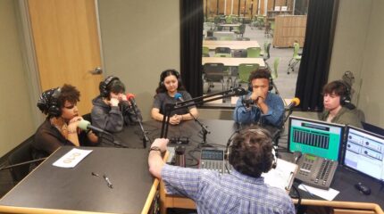 Five teens wearing headphones sitting behind microphones being interviewed by an adult wearing headphones sitting behind a microphone and a mixing board in a radio studio