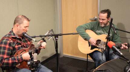 An adult playing mandolin and singing into a microphone and an adult playing acoustic guitar in a radio studio