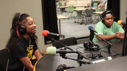 Two teens wearing headphones speaking into microphones in a radio studio