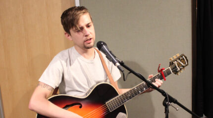 An adult playing acoustic guitar and singing into a microphone in a radio studio