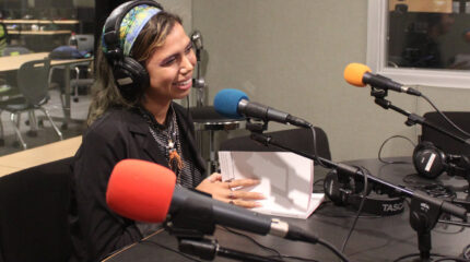 A teen wearing headphones speaking into a microphone and flipping through pieces of paper in a radio studio
