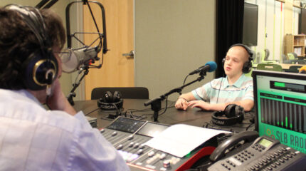 A teen wearing headphones speaking into a microphone being interviewed by an adult wearing headphones sitting behind a microphone and a mixing board in a radio studio