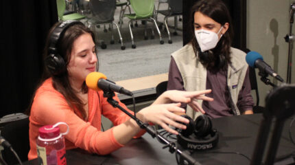 Two teens, one wearing headphones and gesturing with their hands, sitting behind microphones in a radio studio