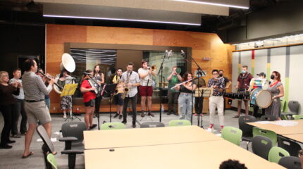 An adult playing clarinet, three adults playing trumpet, an adult playing tuba, an adult playing accordion, two adults playing tenor saxophone, two adults playing baritone saxophone, two adults playing trombone, an adult playing flute, an adult playing cymbals, an adult playing melodica, an adult playing a snare drum, an adult playing a set of toms, and two adults playing bass drum