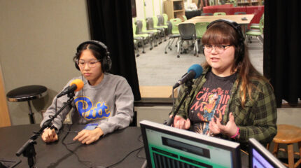 Two teens wearing headphones speaking into microphones in a radio studio