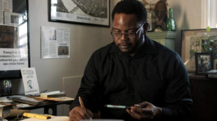 An adult sitting and writing in a book with a pen while holding another pen in their other hand