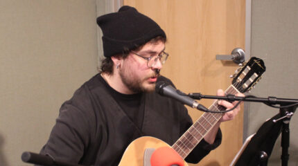 An adult playing an acoustic guitar and singing into a microphone in a radio studio