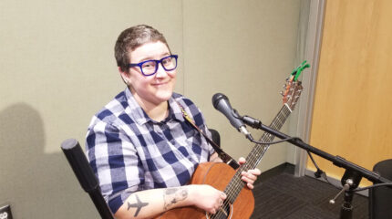 An adult holding an acoustic guitar sitting behind a microphone and smiling at the camera in a radio studio