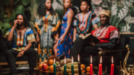 Five adults wearing traditional African clothing sitting in front of Kwanzaa candles and assorted fruits