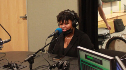 A teen wearing headphones and a face mask smiling while sitting behind a microphone in a radio studio