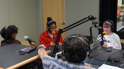 Three teens wearing headphones speaking into microphones being interviewed by an adult wearing headphones sitting behind a microphone and a mixing board in a radio studio
