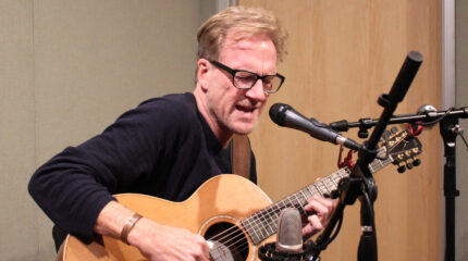 An adult playing acoustic guitar and singing into a microphone in a radio studio