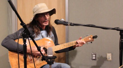 An adult playing acoustic guitar and singing into a microphone in a radio studio