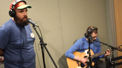 An adult wearing headphones singing into a microphone and an adult wearing headphones playing an acoustic guitar in a radio studio