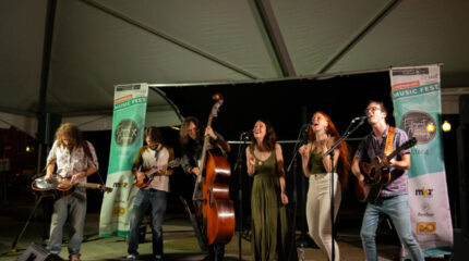 An adult playing a dobro, an adult playing a mandolin, an adult playing an upright bass, two adults singing into microphones, and an adult playing acoustic guitar and singing into a microphone