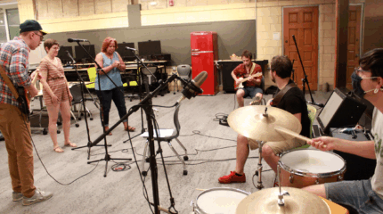 Two adults playing electric guitar, two adults standing behind microphones, an adult playing electric bass, and an adult playing a drum set