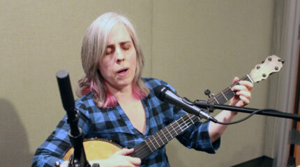 An adult singing into a microphone and playing acoustic guitar in a radio studio