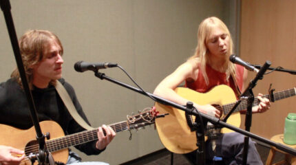 Two adults playing acoustic guitar and singing into microphones in a radio studio