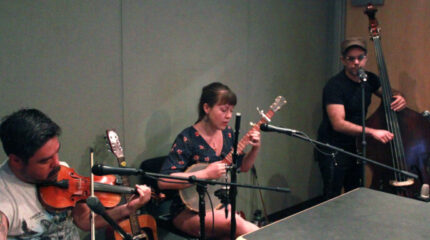 An adult playing violing into a microphone, an adult playing banjo, and an adult playing upright bass in a radio studio