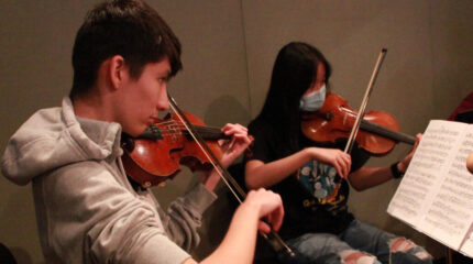 Two teens playing violins looking at sheet music on music stands in a radio studio