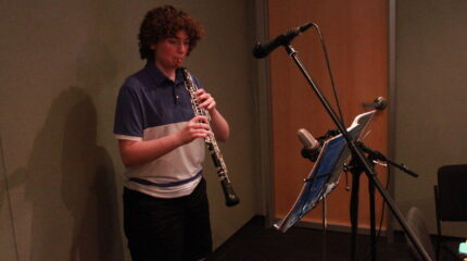 A teen playing oboe into a microphone looking at sheet music on a music stand in a radio studio