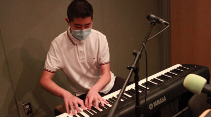 A teen playing a piano keyboard sitting behind a microphone in a radio studio