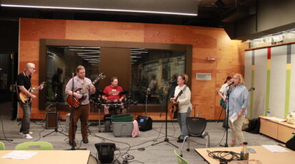 Two adults playing electric guitar, an adult playing an electric drumset, an adult playing an acoustic guitar and singing into a microphone, an adult playing electric bass, and an adult singing into a microphone