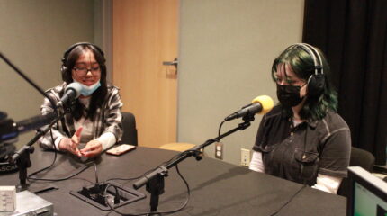 Two teens wearing headphones and face masks speaking into microphones in a radio studio