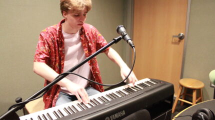 A teen playing a piano keyboard sitting behind a microphone in a radio studio