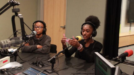 Two teens wearing headphones, one gesturing with their hands and speaking into a microphone in a radio studio