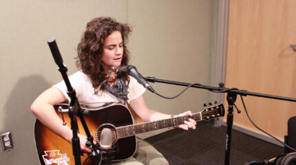 An adult singing into a microphone and playing acoustic guitar in a radio studio