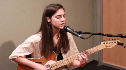 An adult playing acoustic guitar and singing into a microphone in a radio studio