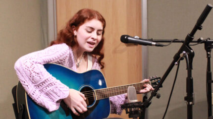 An adult playing acoustic guitar and singing into a microphone in a radio studio