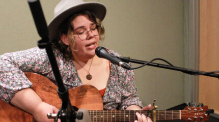 An adult playing acoustic guitar and singing into a microphone in a radio studio