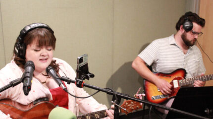 Two adults wearing headphones, one playing electric guitar and one playing acoustic guitar and singing into a microphone in a radio studio