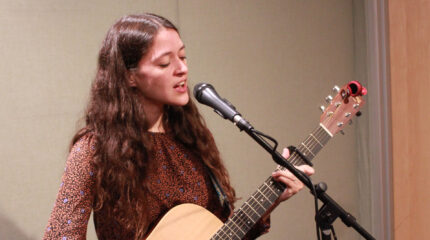 An adult playing acoustic guitar and singing into a microphone in a radio studio