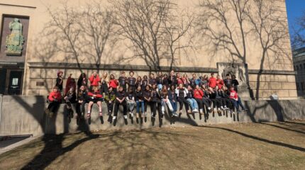 A large group of youth sitting on a wall in front of a large beige building