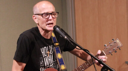 An adult playing acoustic guitar and singing into a microphone in a radio studio