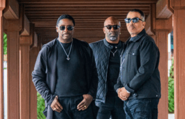 Three adults wearing sunglasses posing under a bridge