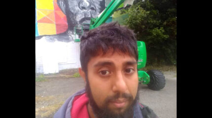 An adult posing in front of an art piece on a wall and a green construction vehicle