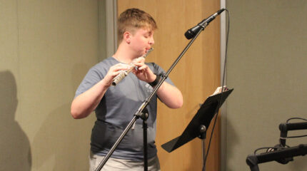 A teen playing a flute into a microphone standing in front of a music stand in a radio studio