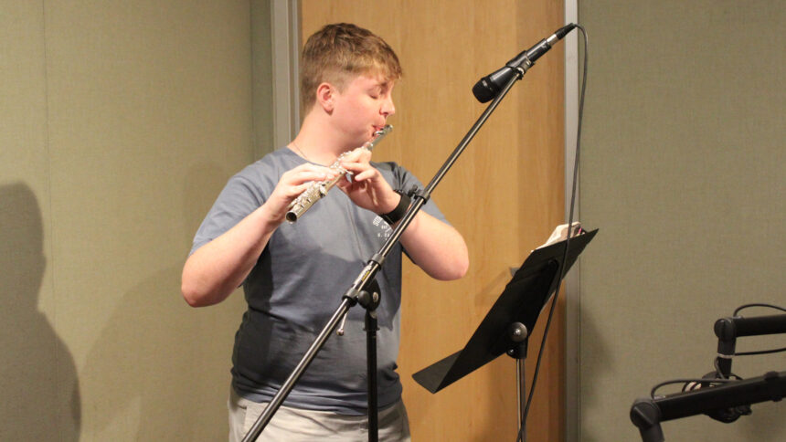 A teen playing a flute into a microphone standing in front of a music stand in a radio studio